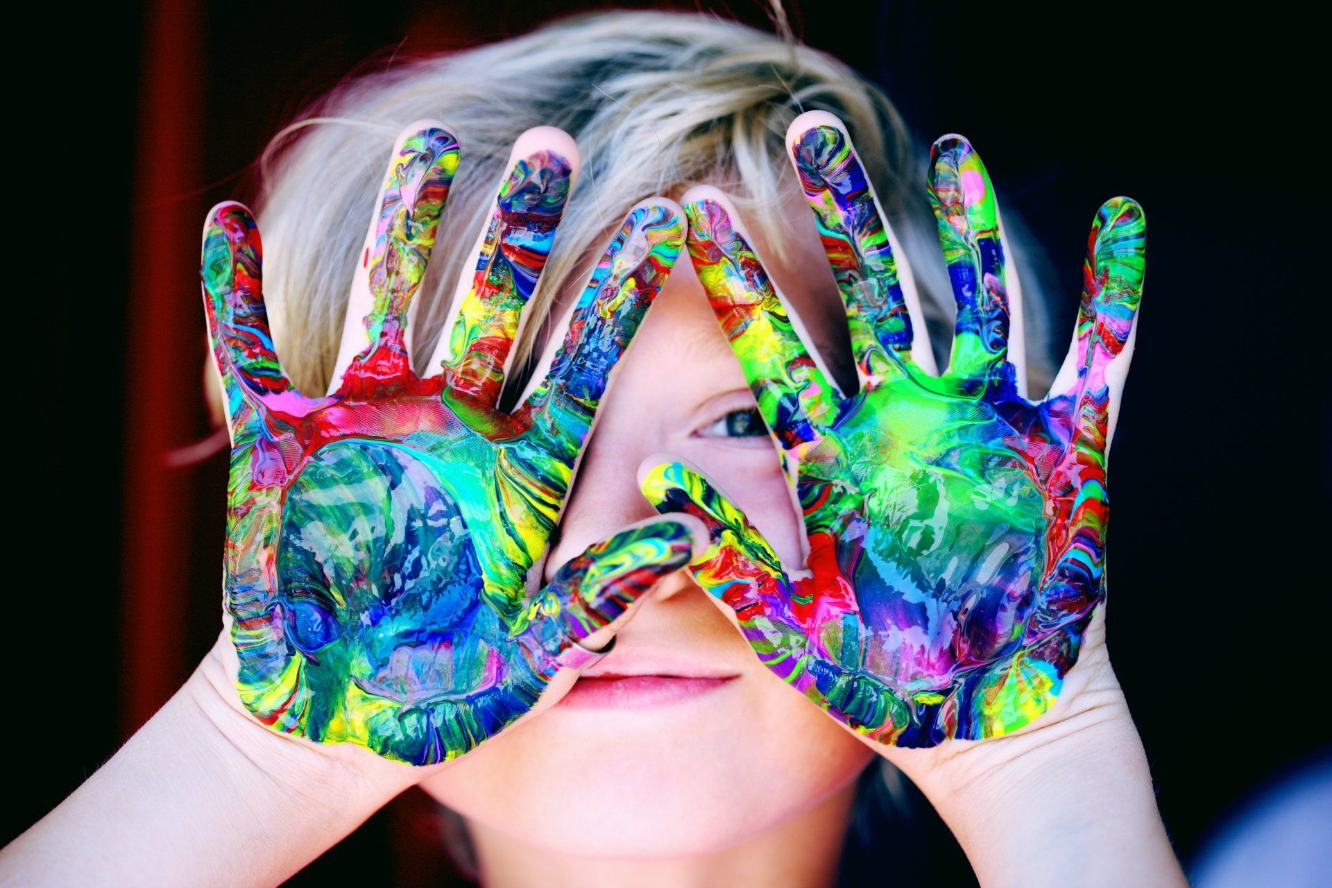 Child with hands covered in colorful paint, looking through them at the viewer.