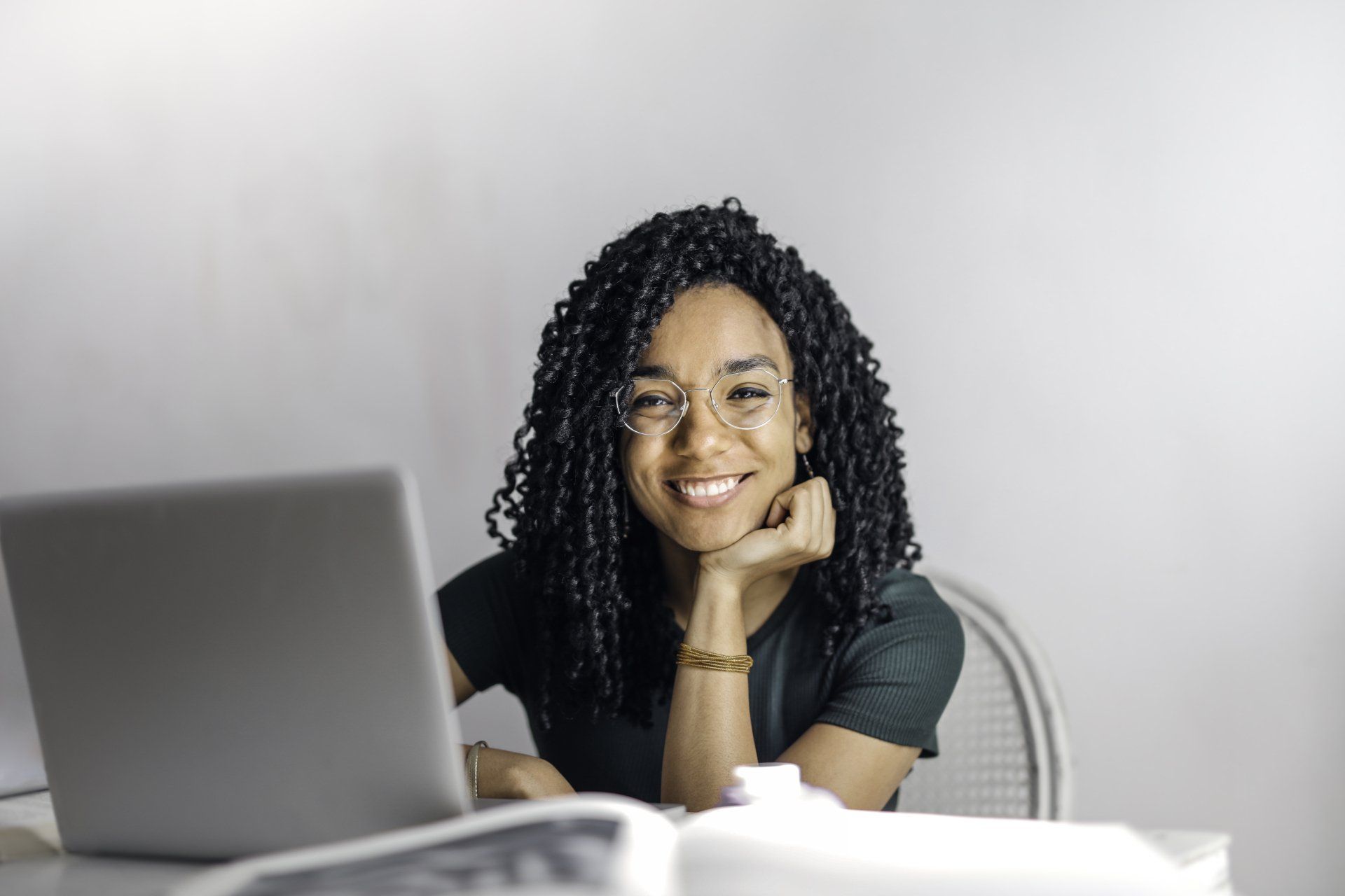 Woman with glasses smiling, leaning on hand at desk with laptop and open book.