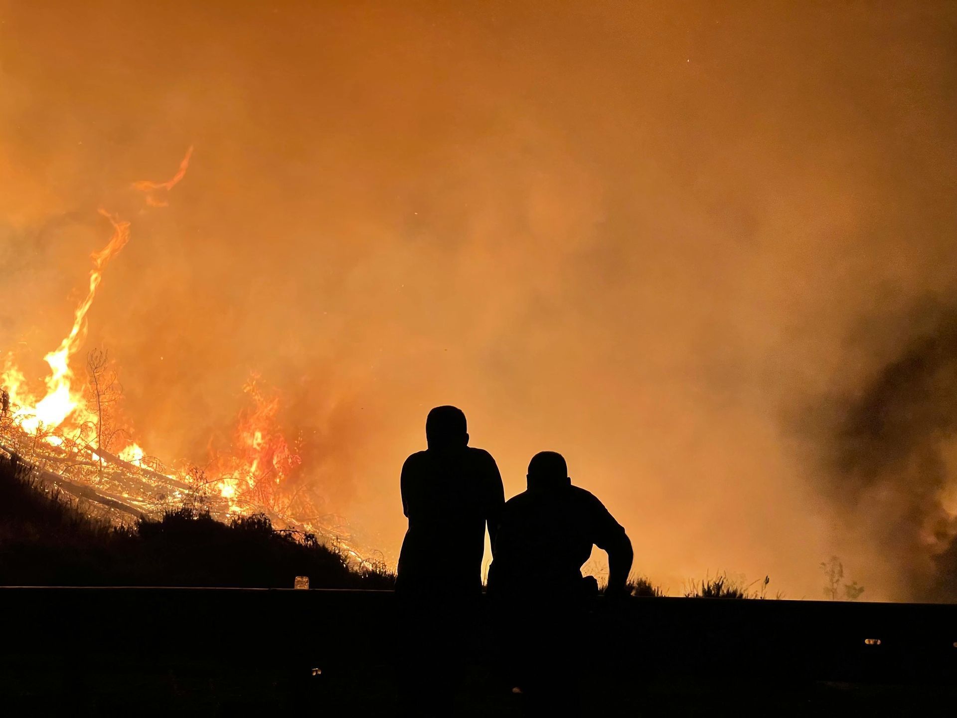 Silhouetted figures watching a wildfire blaze, flames and smoke filling the sky.