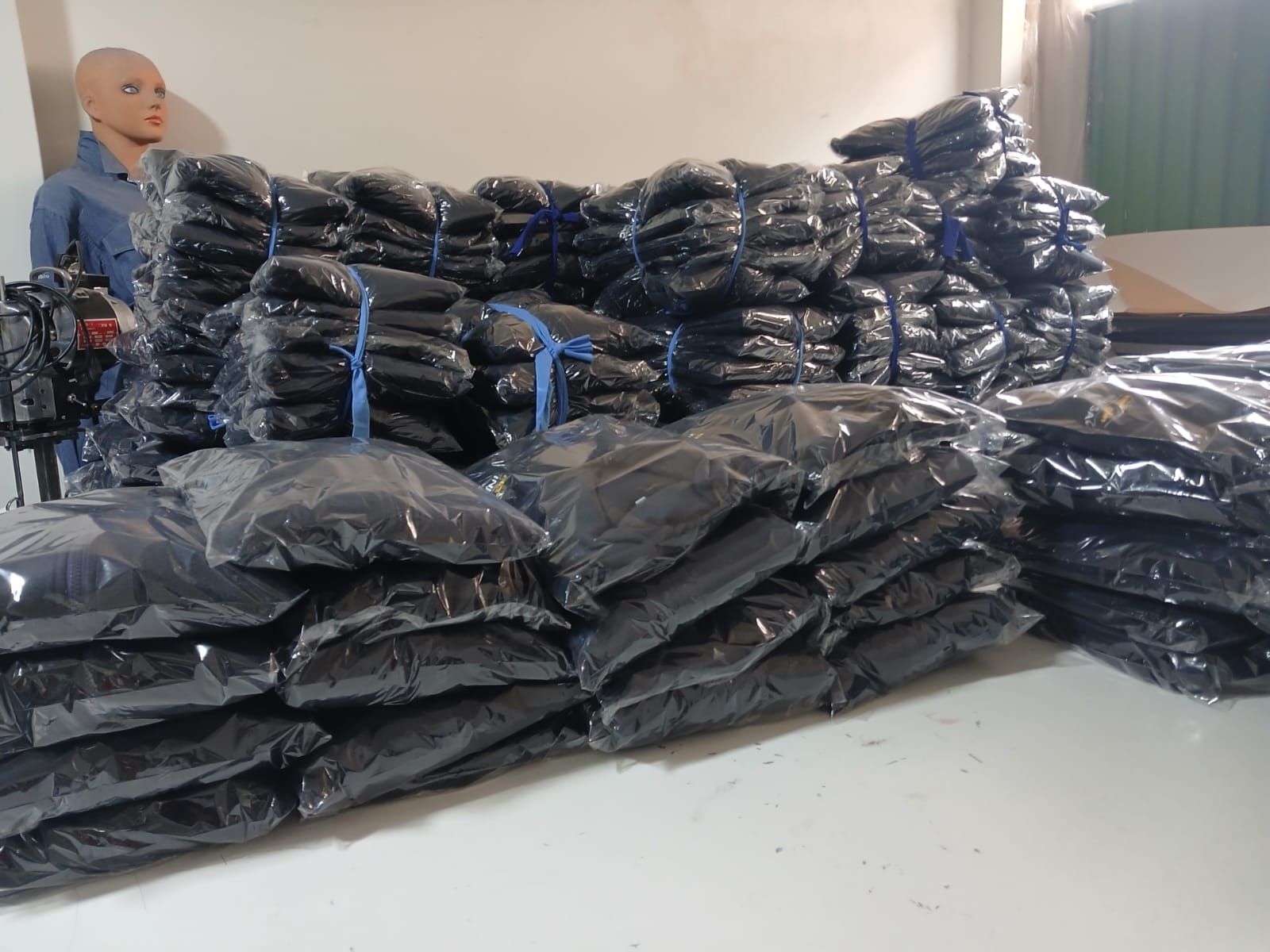 Montones de bolsas negras envueltas en plástico apiladas en una habitación, con una muñeca visible a la izquierda.