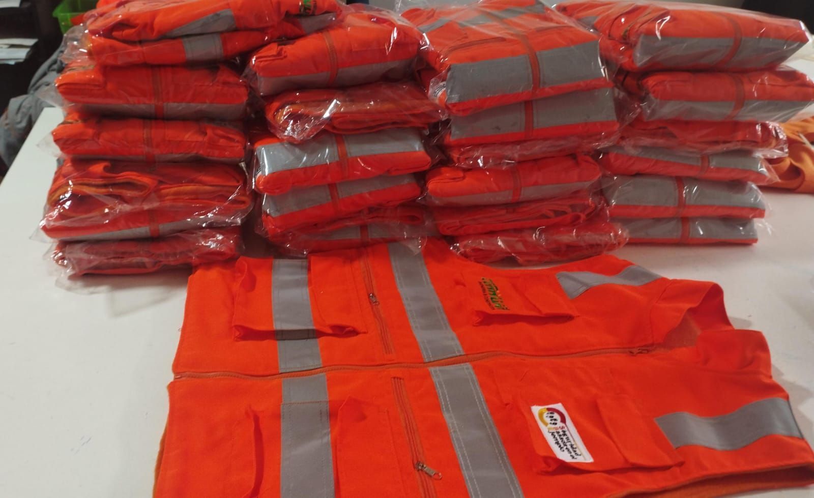 Chalecos de seguridad rojos apilados en envases de plástico con un chaleco reflectante naranja en primer plano.