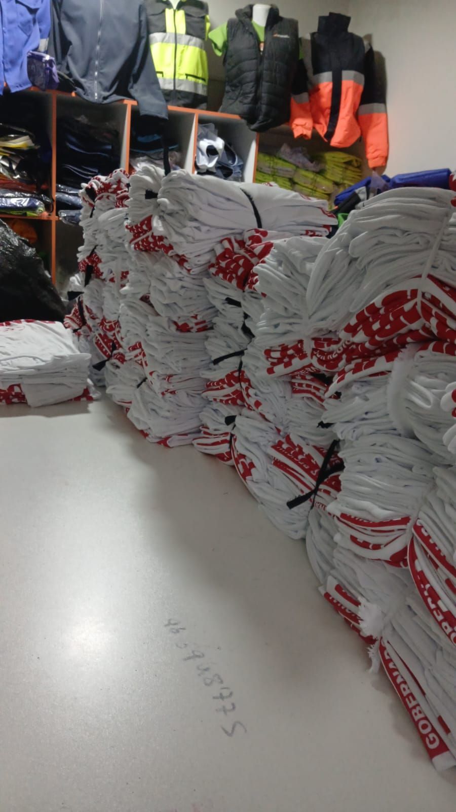 Montones de artículos empaquetados en blanco y rojo en un almacén, con chaquetas y equipos de seguridad colgados al fondo.