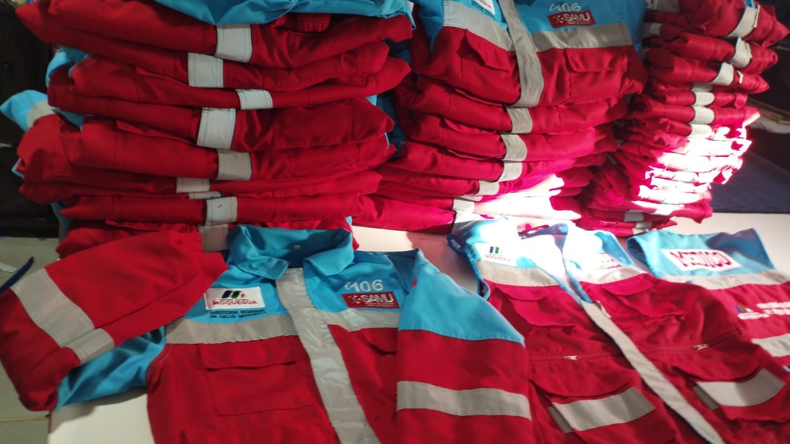 Montones de camisas o uniformes a rayas rojas y azules apilados en un almacén, con la luz del sol a la derecha.