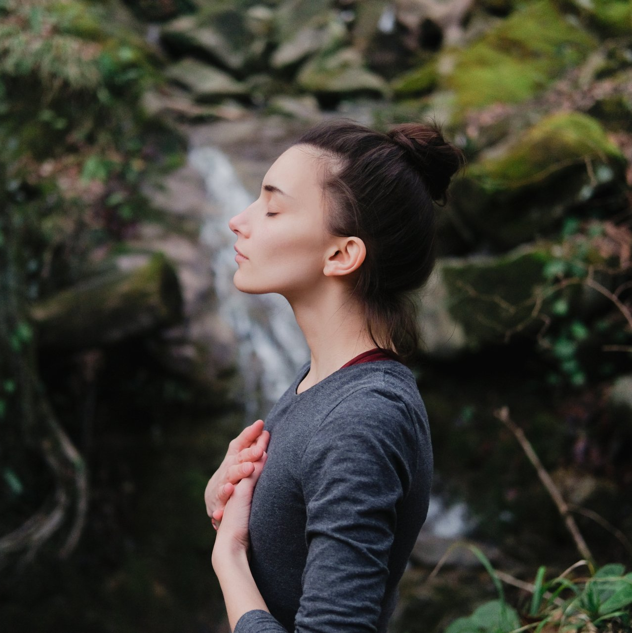 Woman with eyes closed, hands on chest, by a small waterfall, in nature.