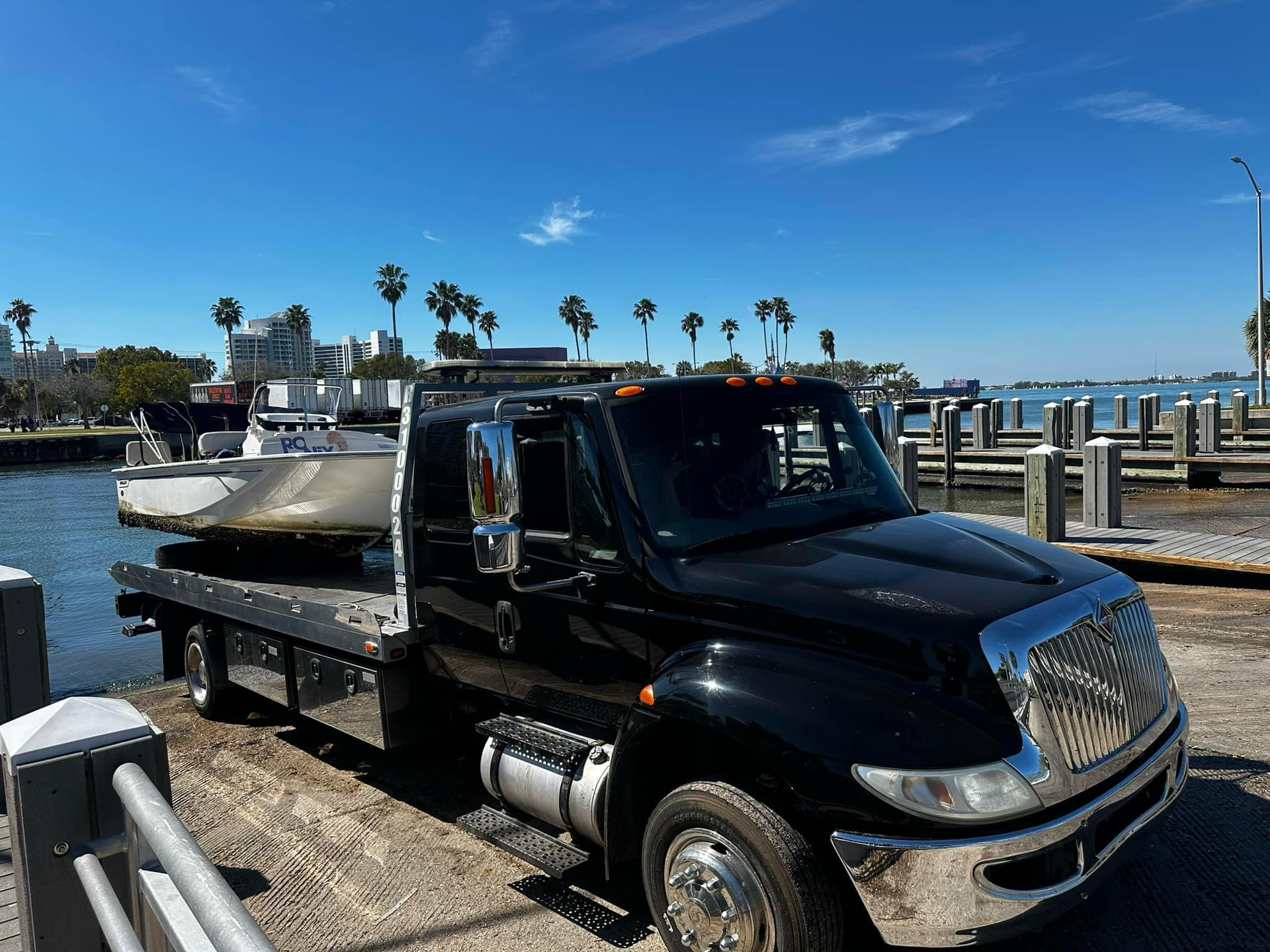 A black tow truck with a boat on the back is parked next to a body of water.