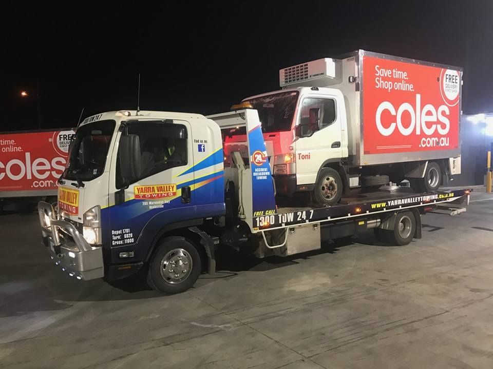 A Coles delivery truck being transported on a tow truck at night. Both trucks are white and have Coles branding.