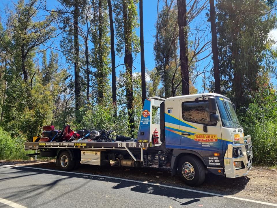 Tow truck parked on a roadside, transporting motorcycles. The truck is blue and white, set in a forested area.
