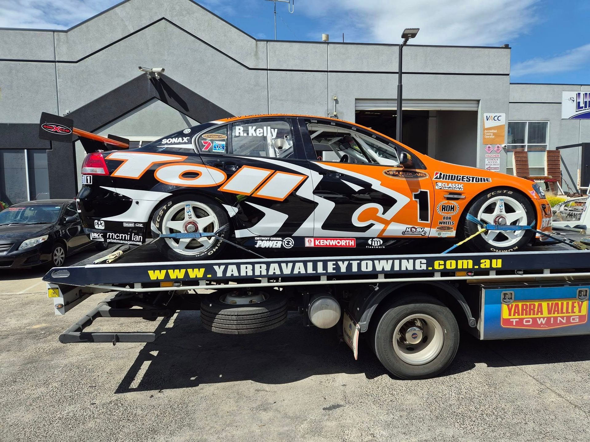 A race car, with the Toll logo, is secured on a tow truck in front of a building. The car is black, orange, and white.