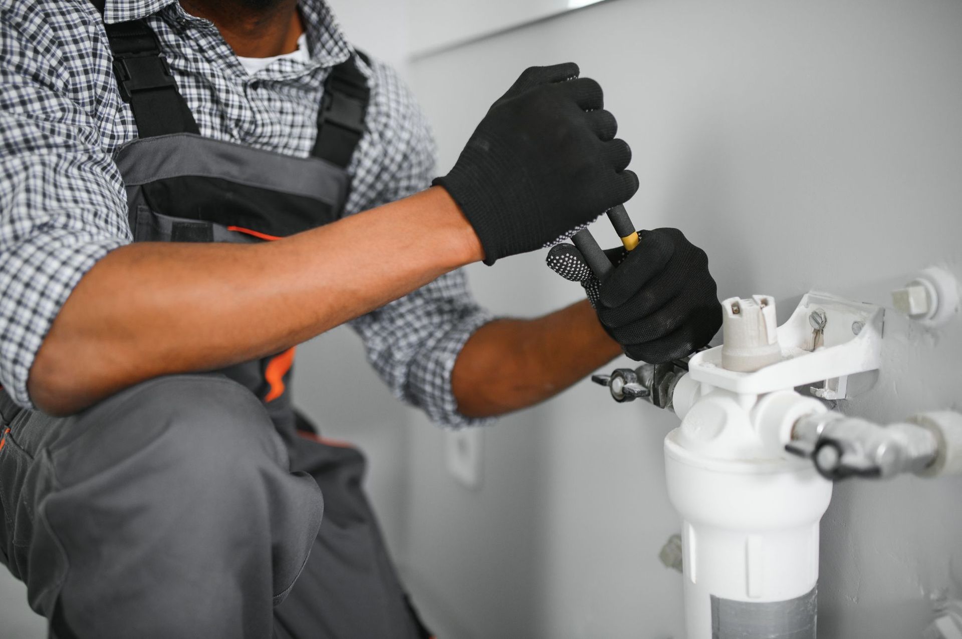 Plumber using a wrench on a white water filter system near a wall, wearing gloves and overalls.