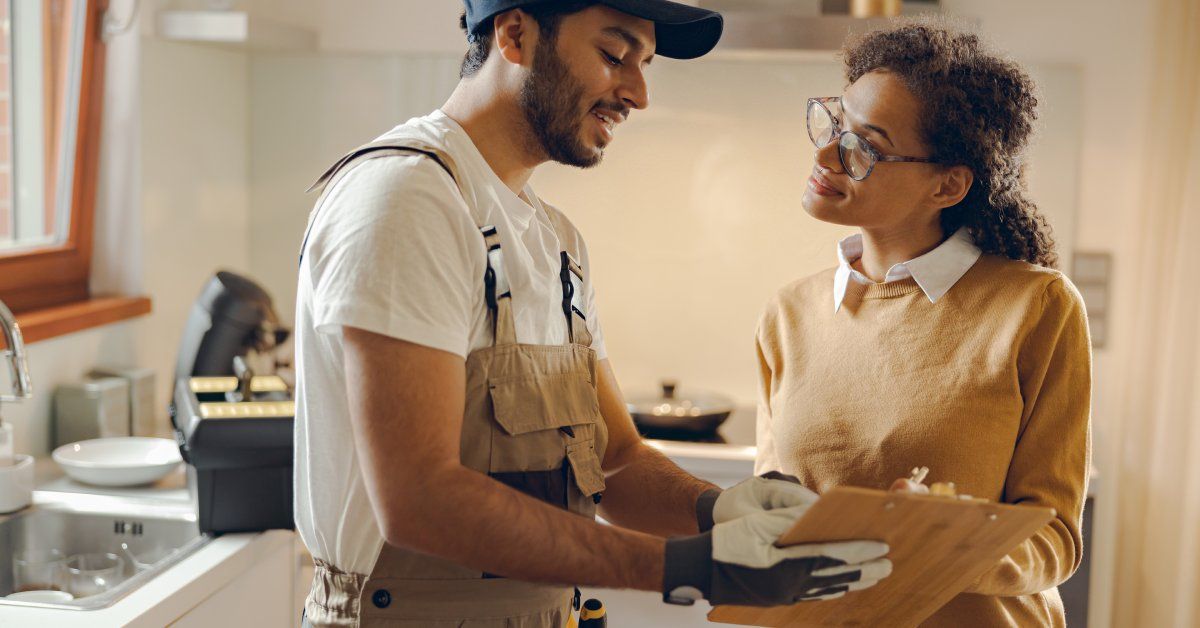 A young handyman in coveralls and gloves, up close, showing a clipboard to a female client in a kitchen.