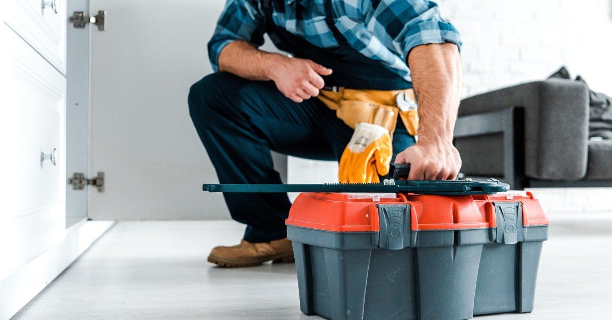  A handyman in a plaid shirt and overalls, up close, holding a toolbox as he crouches to look inside of a cabinet.