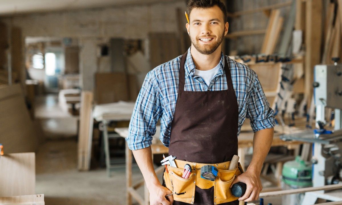 A young handyman with a flannel shirt, apron, and tool belt stands in his workshop smiling.