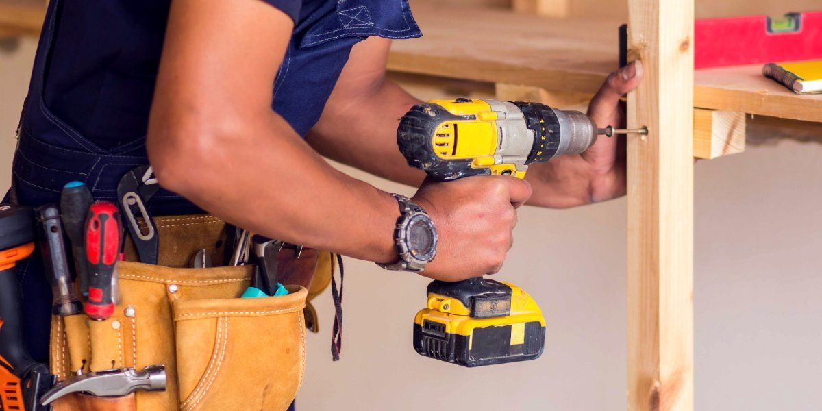 A handyman wearing a watch and a toolbelt using a drill to place a screw into a piece of plywood.