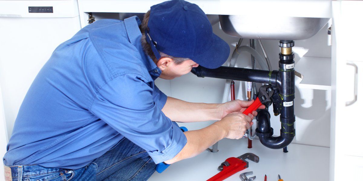 A handyman in jeans, a blue shirt, and hat uses a large wrench to fix the plumbing under a kitchen sink.