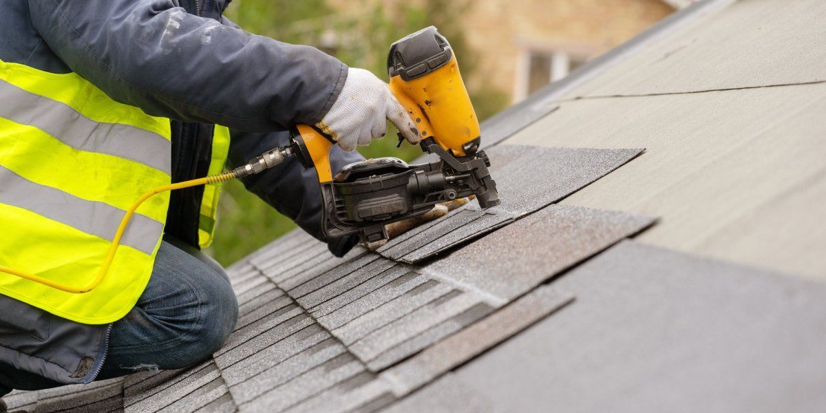 A man in a yellow high-visibility vest using a pneumatic nail gun to install roofing shingles.