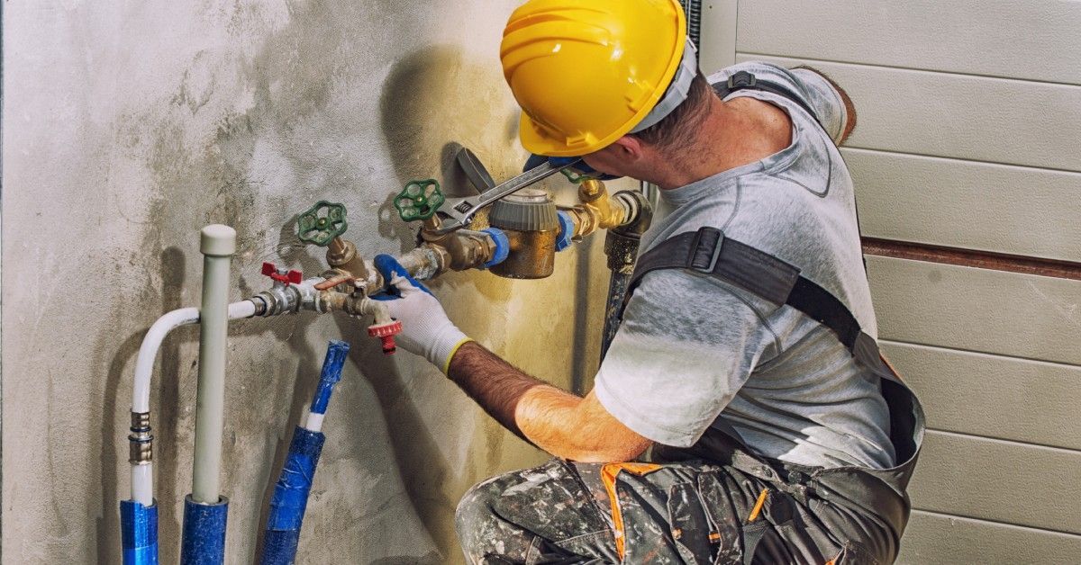 A professional handyman wears overalls and a yellow helmet. He kneels next to the basement's plumbing work.
