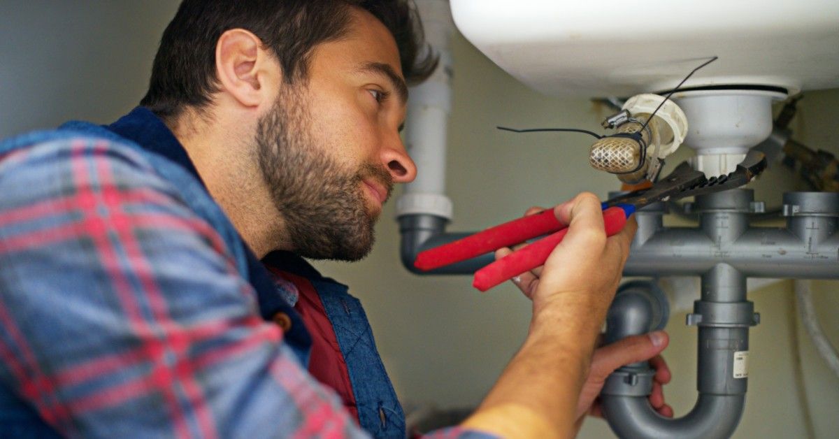 A professional handyman wears a blue and red flannel shirt as he works under the sink.