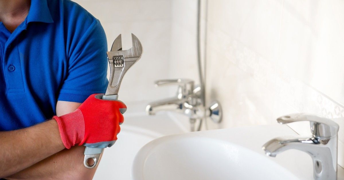 A professional handyman wears a blue work shirt and holds a large wrench with a red glove on. The bathroom is behind him.
