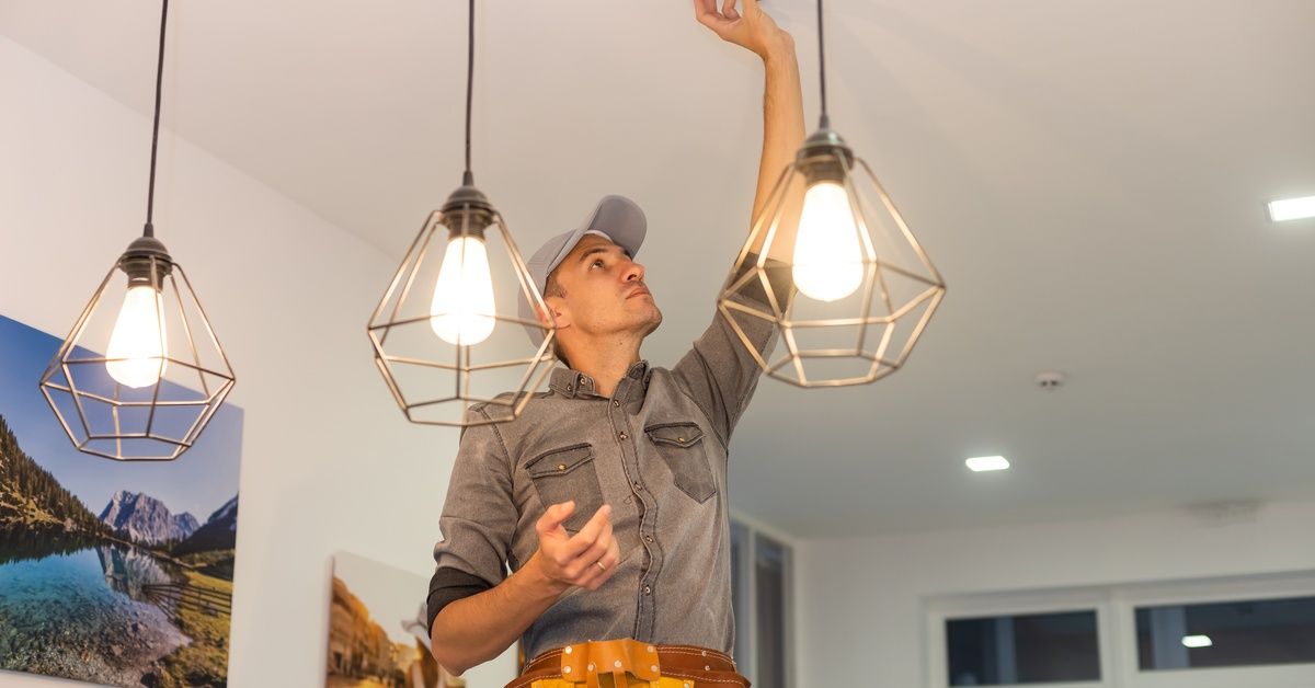 A professional handyman stands on a stool near the kitchen island as he adjusts the smoke detector and wears a tool belt.