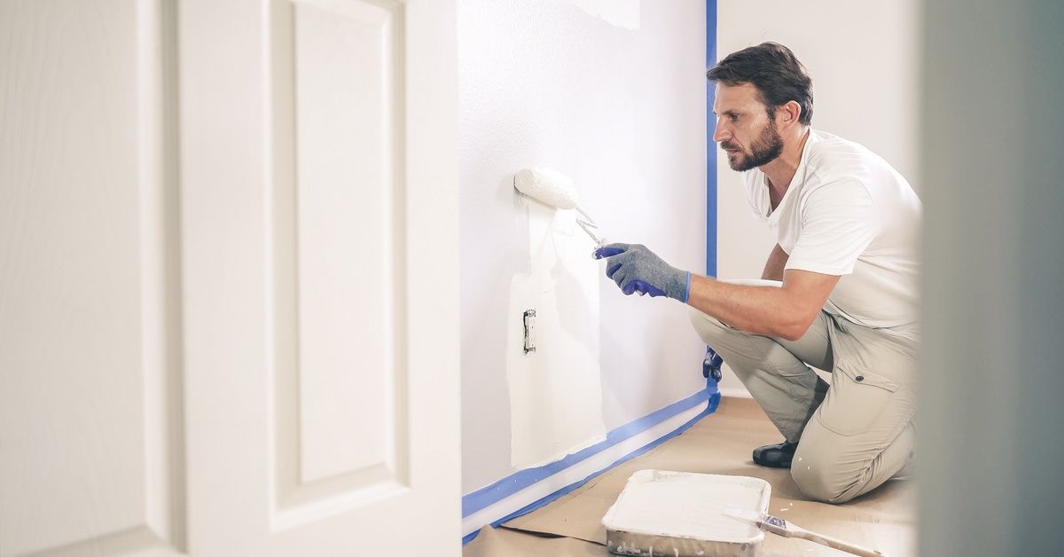 A professional handyman kneels down in painting gear as he rolls the walls with a paint roller full 