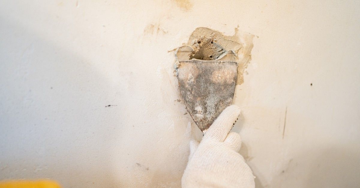 A handyperson wears a white glove as they patch a hole in the drywall with plaster. The plaster is gray.
