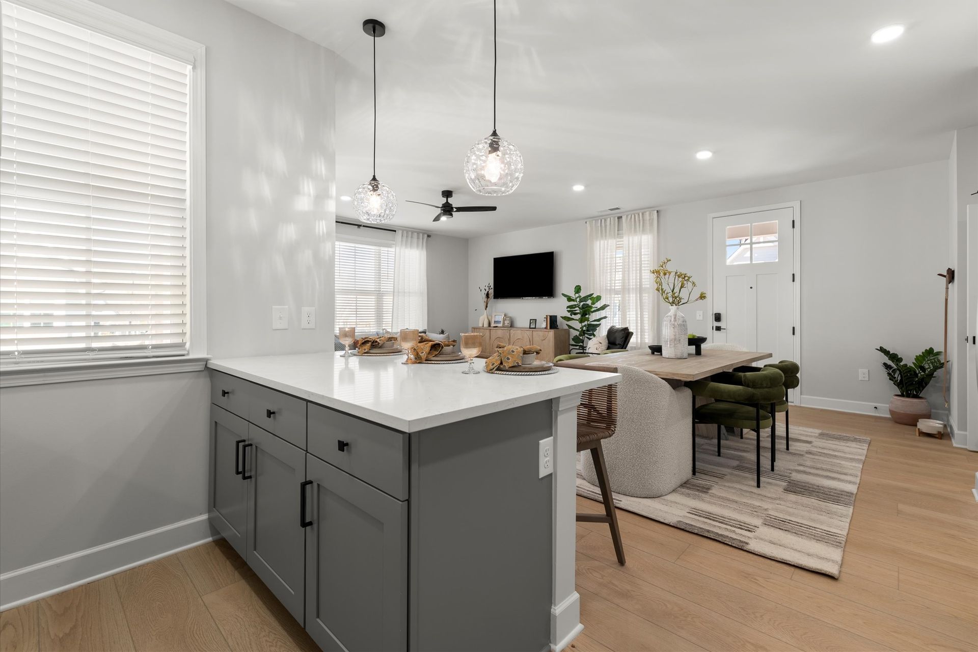 Open-concept kitchen and living area with gray island, dining table, and television; natural light, light wood floors.