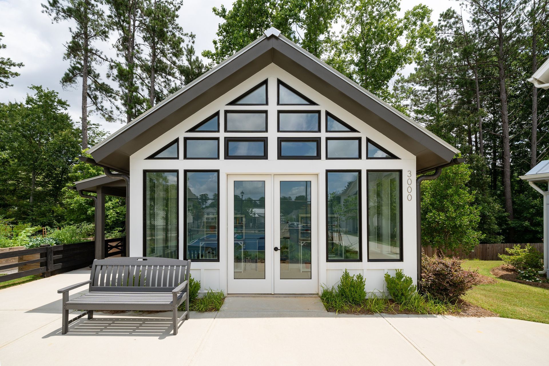 White modern building with glass windows and doors, gray roof, and wooden bench on a concrete patio, surrounded by trees.