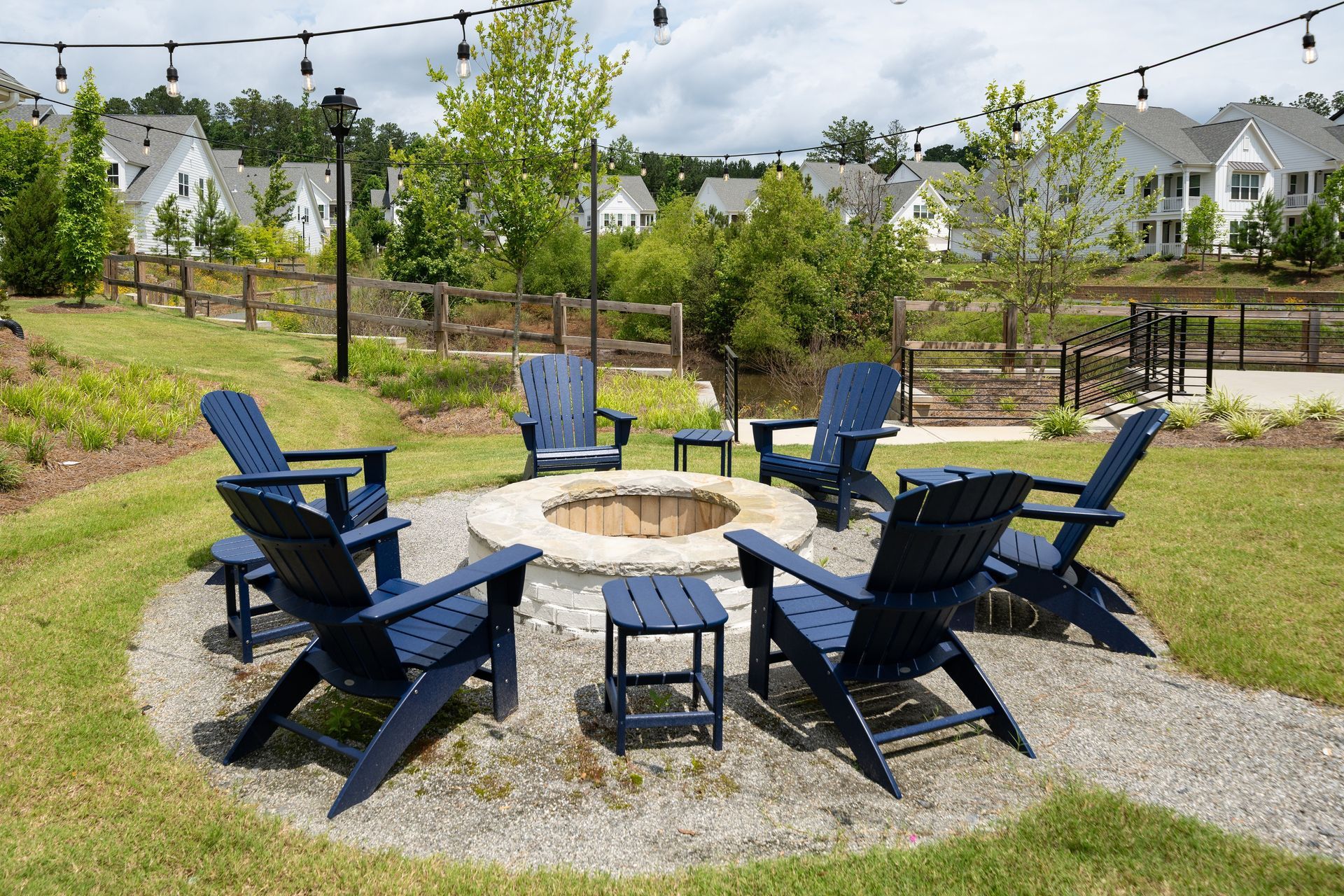A fire pit surrounded by blue Adirondack chairs in a grassy area with string lights and houses in the background.
