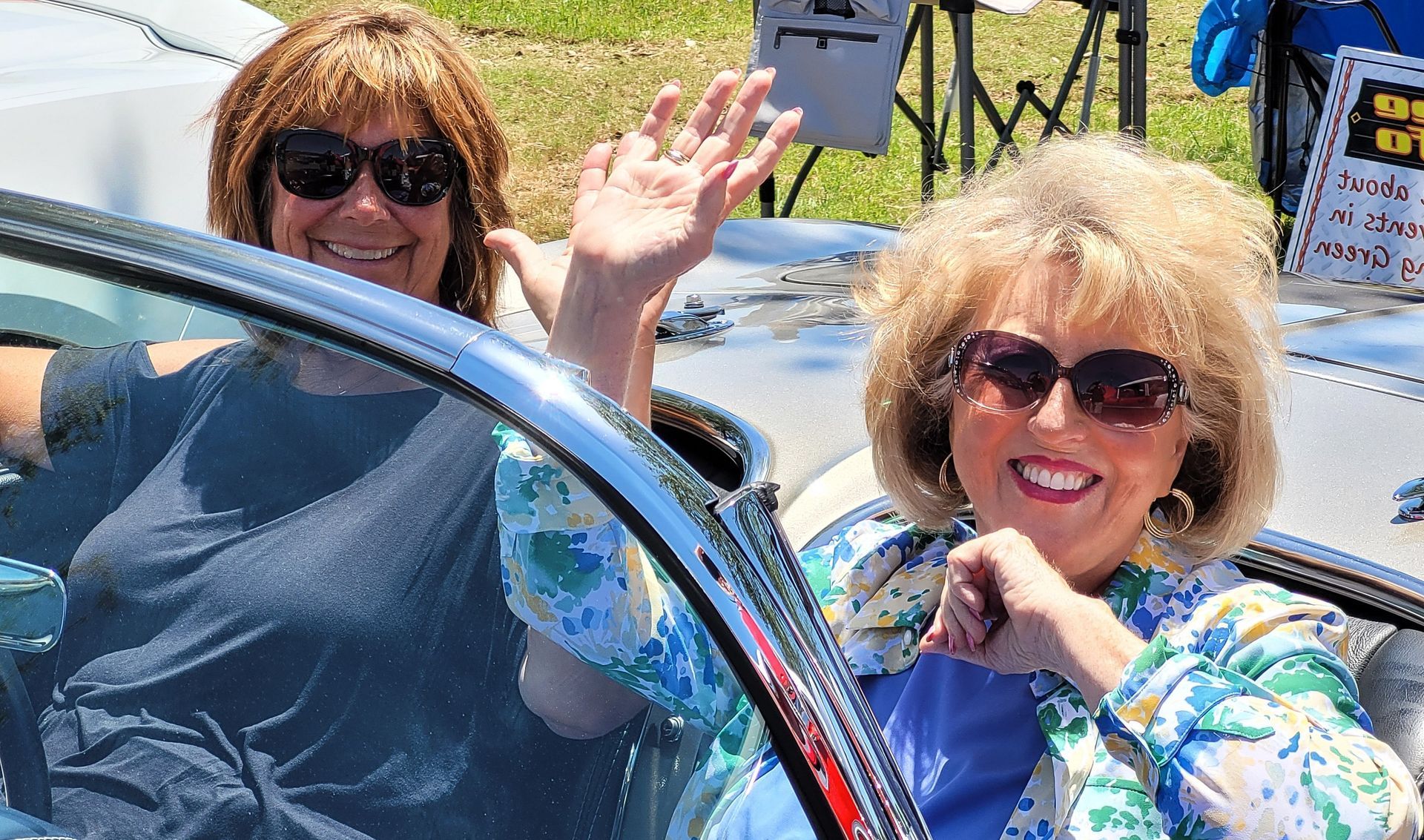 Two smiling women in sunglasses wave from a white convertible car on a sunny day.
