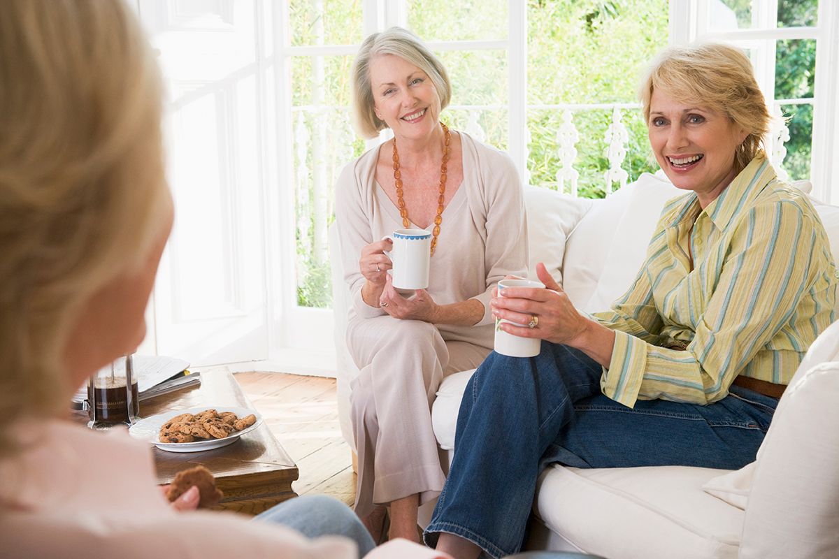 Three smiling senior women in a living room, one facing away, two holding mugs on a white couch.