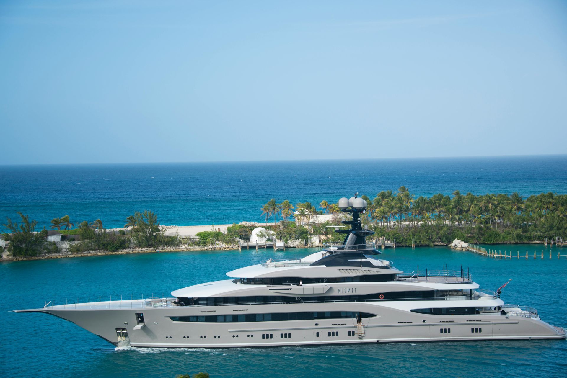 Large, gray luxury yacht sailing in turquoise water near an island with palm trees and blue sky.