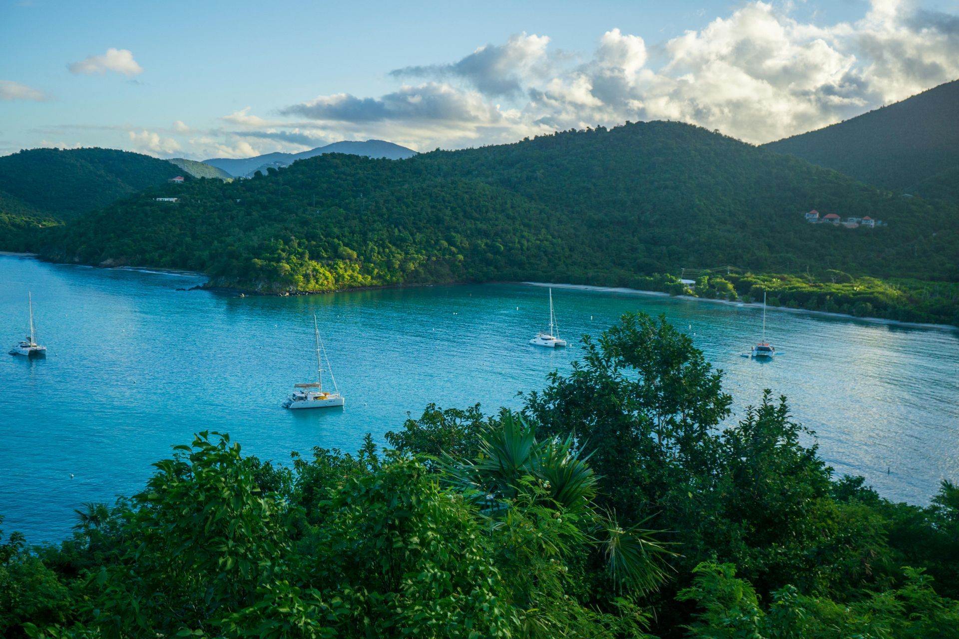 Bay with sailboats, surrounded by lush green hills, under a soft, sunny sky.