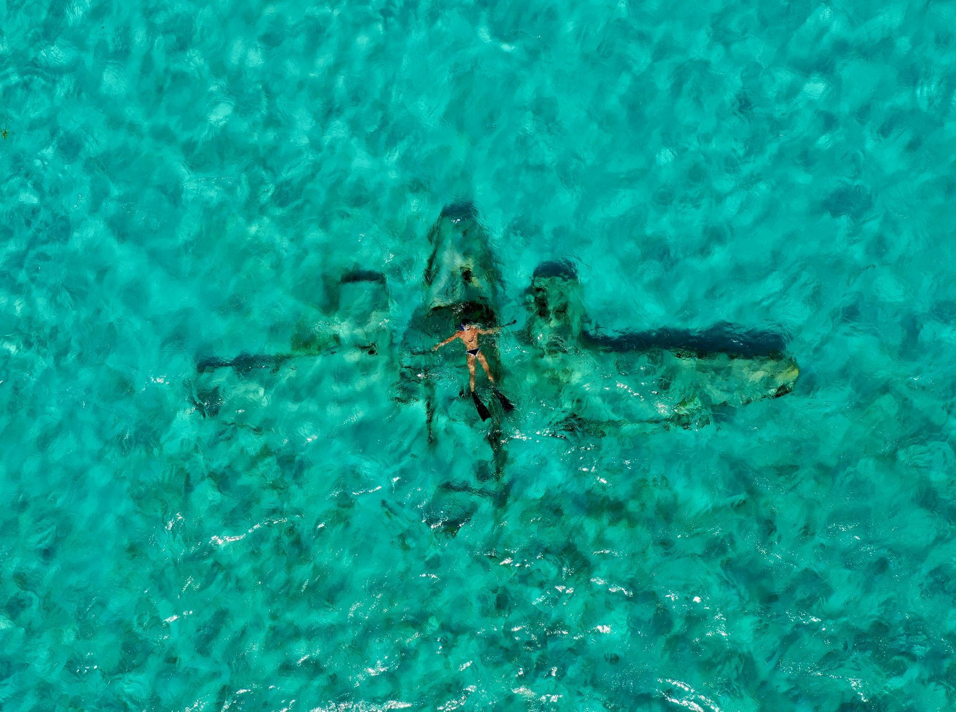 Snorkeler swims over a submerged airplane in clear, turquoise water.
