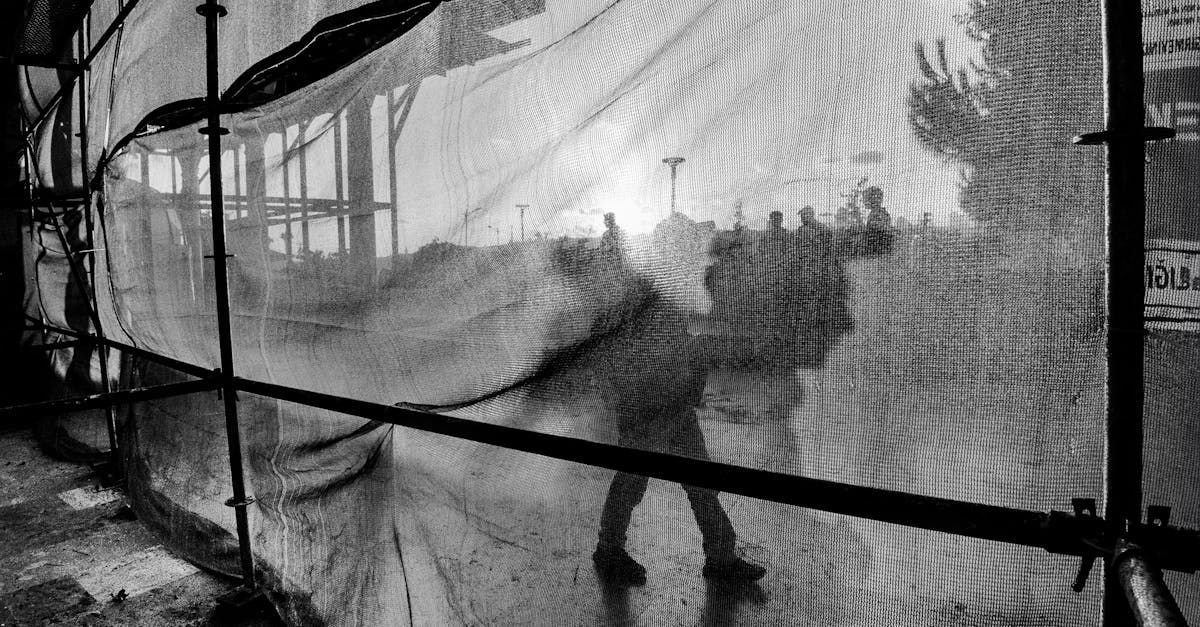 People walking past a construction site, viewed through netting. Black and white.