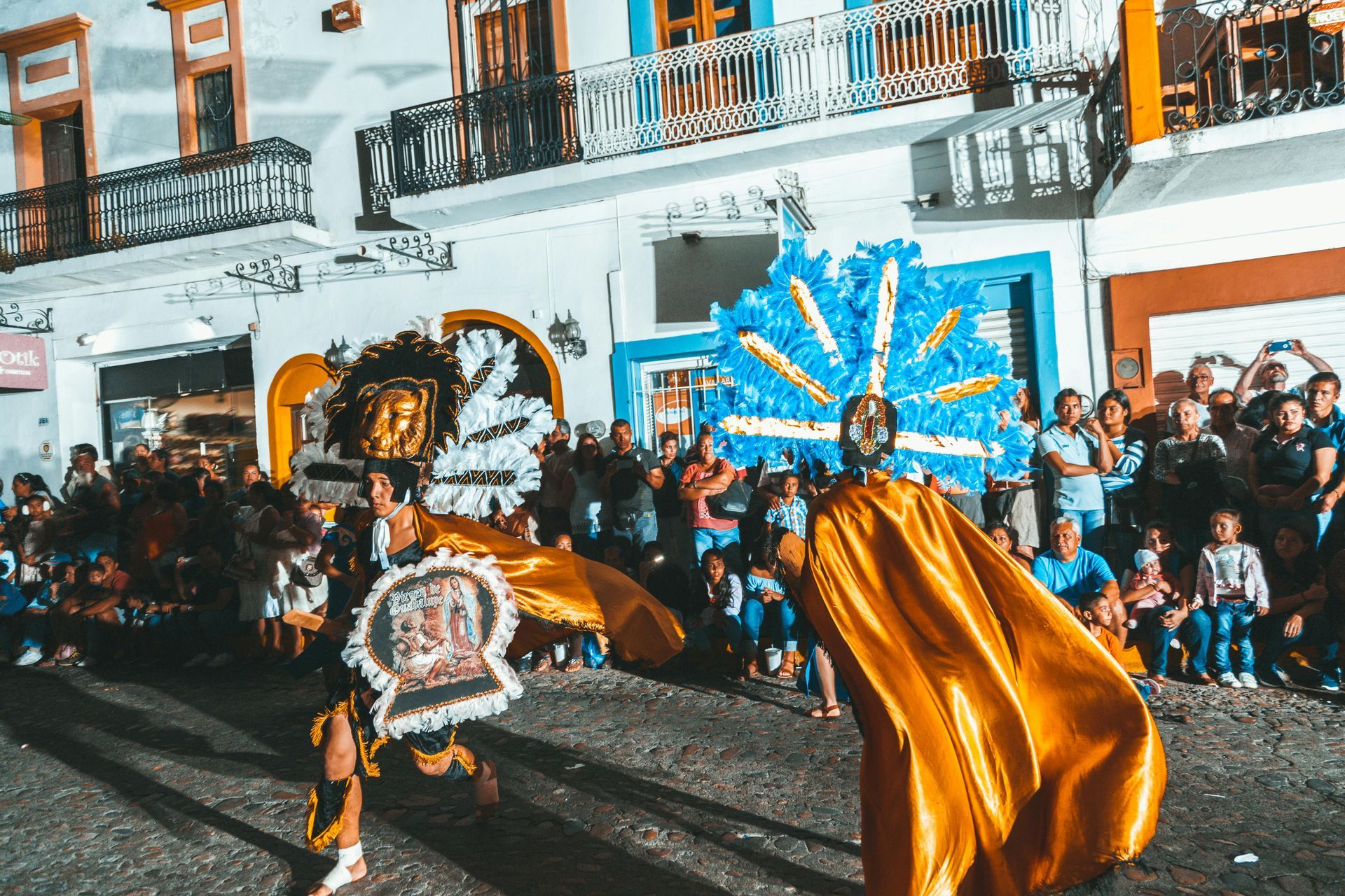 Performers in elaborate costumes dance on a street, with a crowd of people watching against a backdrop of colorful buildings.