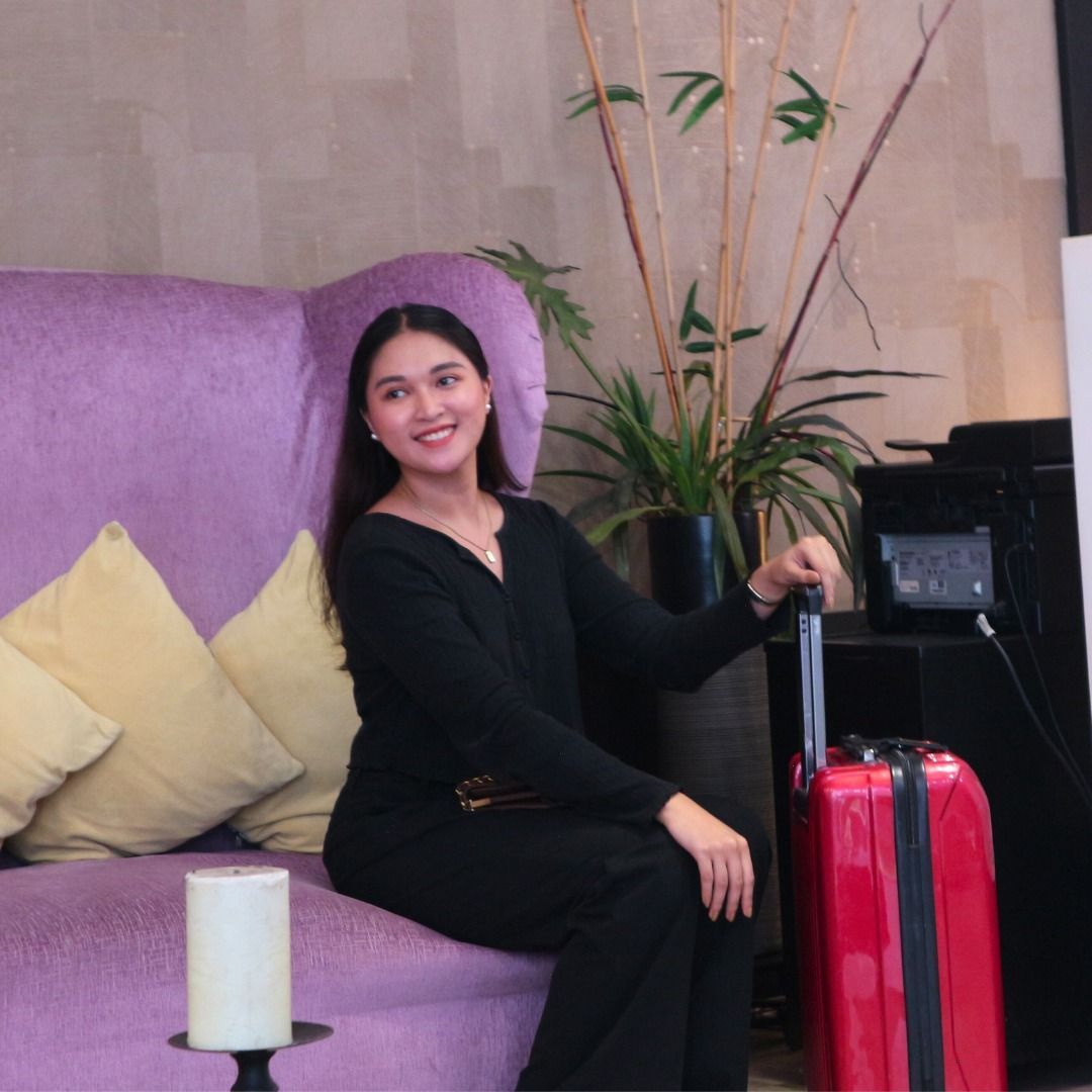 Woman in black outfit smiles while sitting next to red suitcase on a purple couch.