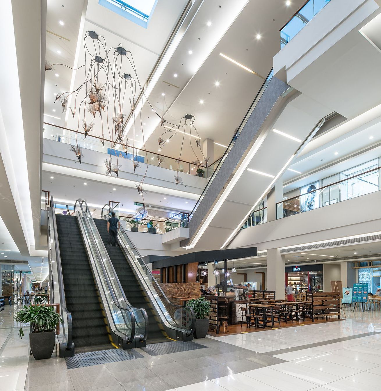 Mall interior with escalators, multiple levels, and shops. A person rides the escalator.