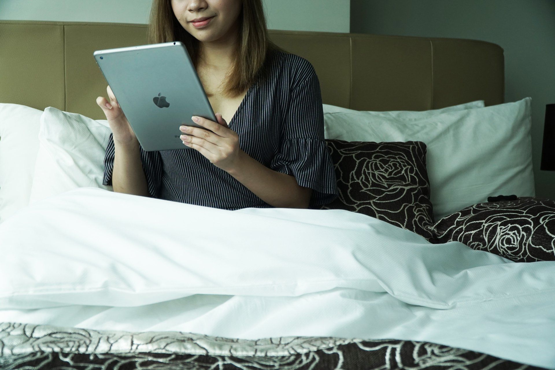 A person sits up in bed against pillows, holding and looking at a tablet.
