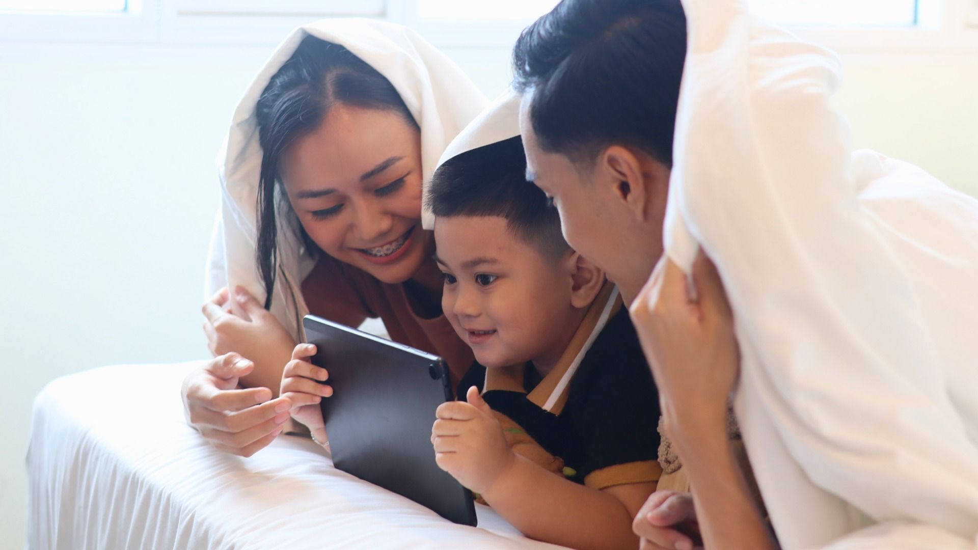 A family is laying under a blanket on a bed looking at a tablet.
