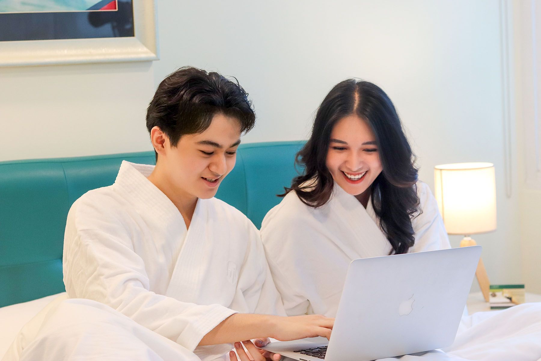 Couple in bathrobes on bed, smiling at laptop screen, hotel room setting.