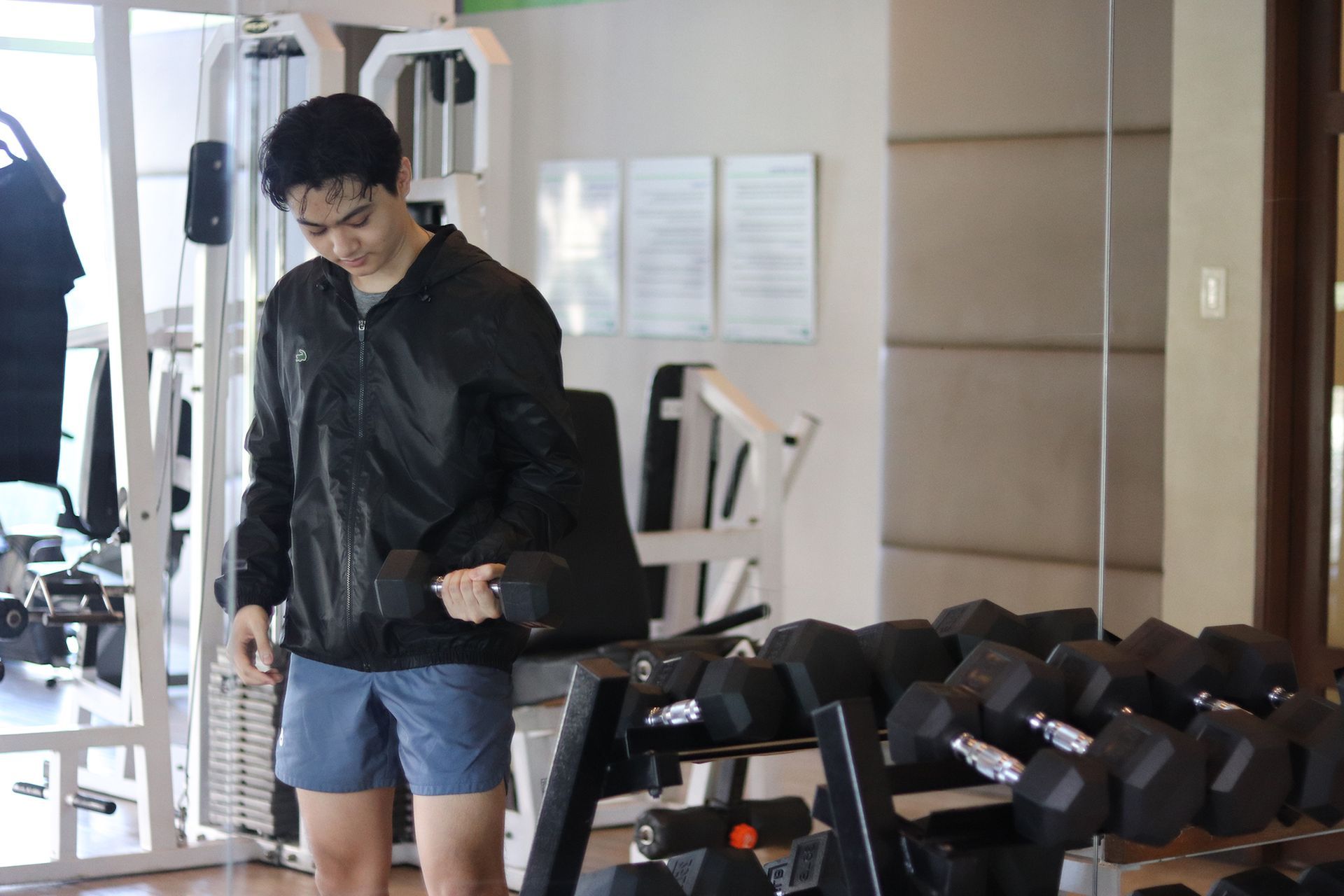 Man in gym lifting a dumbbell, wearing black jacket and blue shorts. Mirrors and weight rack visible.