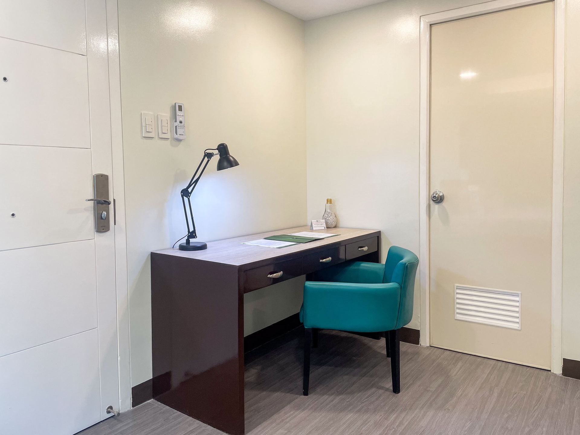 A desk with a lamp and teal chair next to a closed door in a room with light-colored walls.
