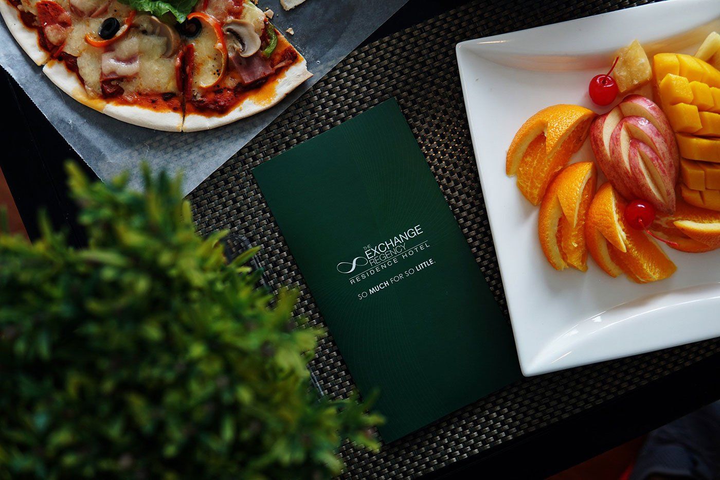 A pizza and a plate of fruit are on a table next to a menu from The Exchange Regency Hotel in Ortigas