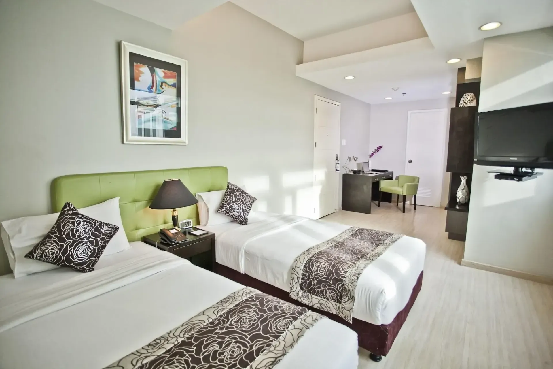 Two twin beds in a hotel room with a desk, TV, and artwork. Green and neutral colors.