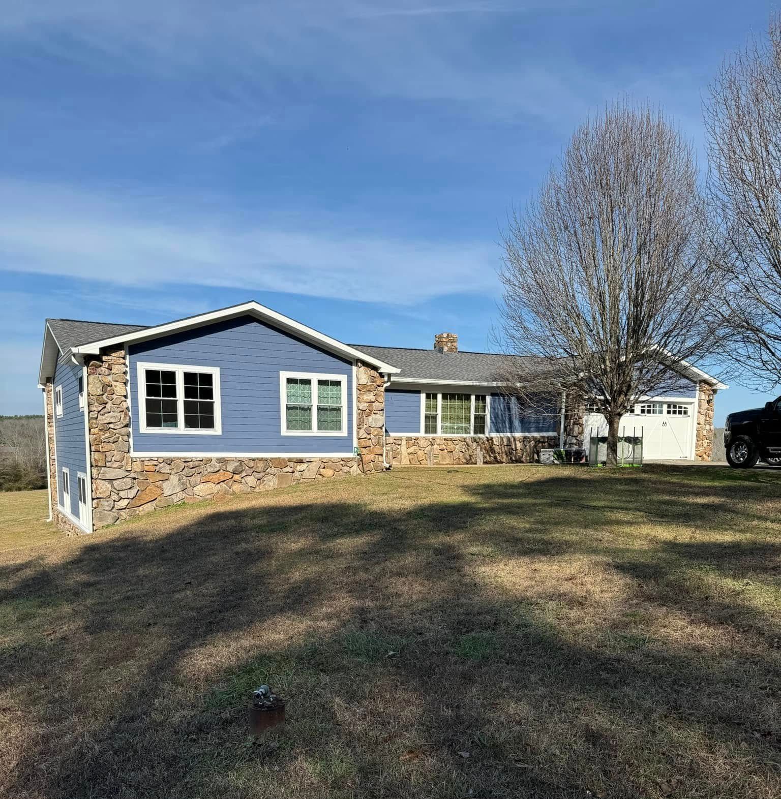 Blue-sided ranch house with stone accents, windows, and a garage door, set on a grassy hill under a blue sky.