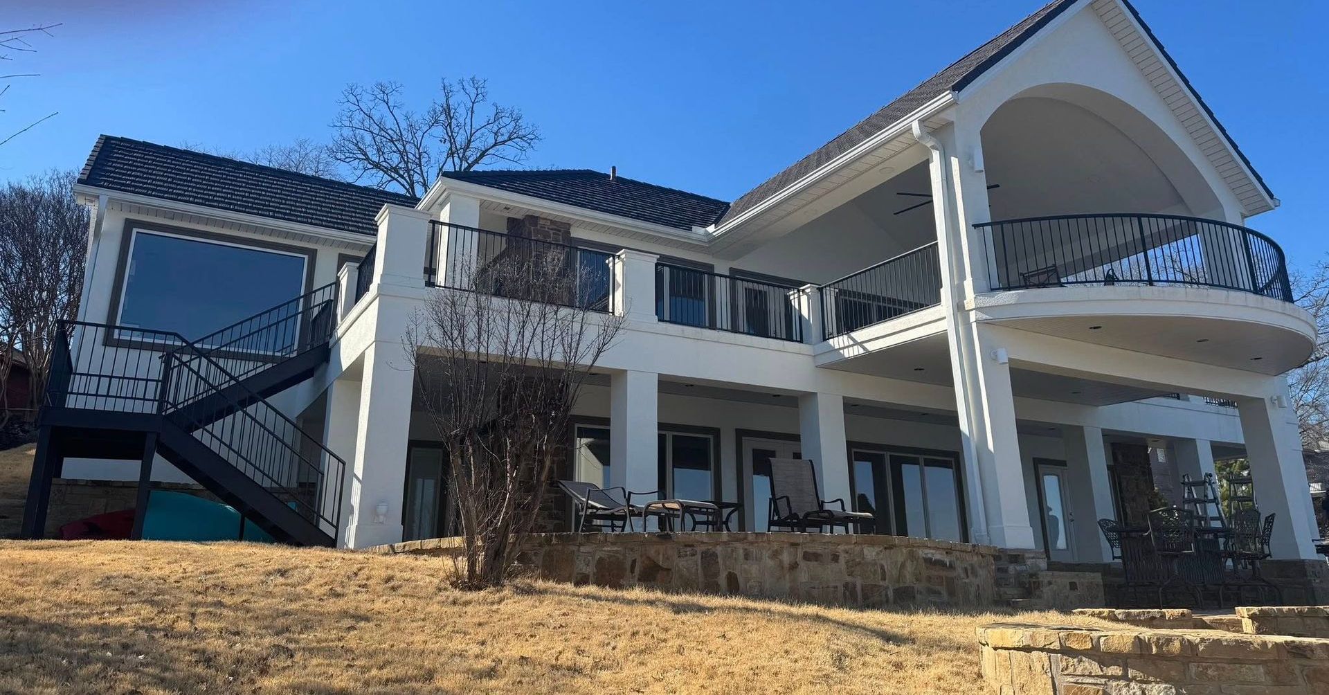 White two-story house with black roof and balconies, set on a hillside against a blue sky.
