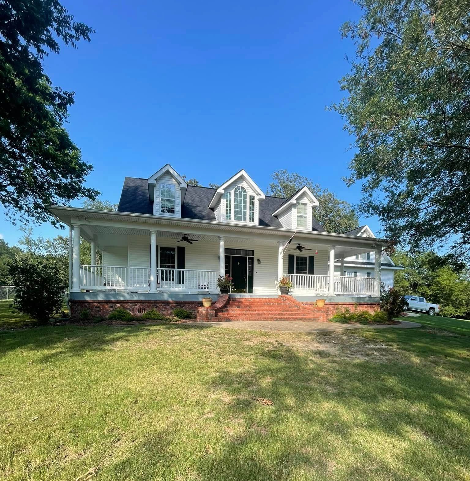 White farmhouse with a wraparound porch and brick foundation under a clear, blue sky.