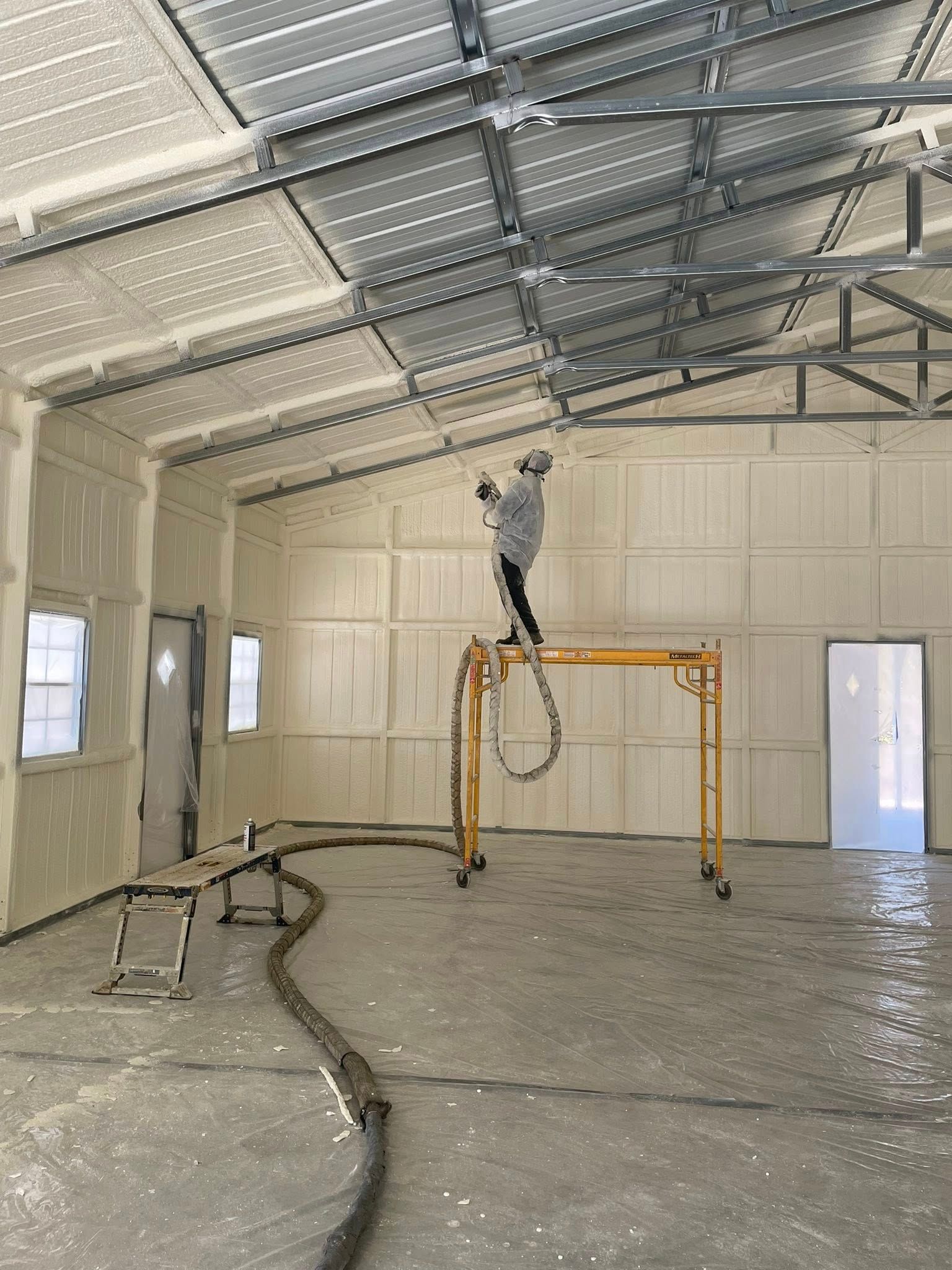 Man spraying insulation inside a building; walls and roof. Worker on scaffolding, hose visible.