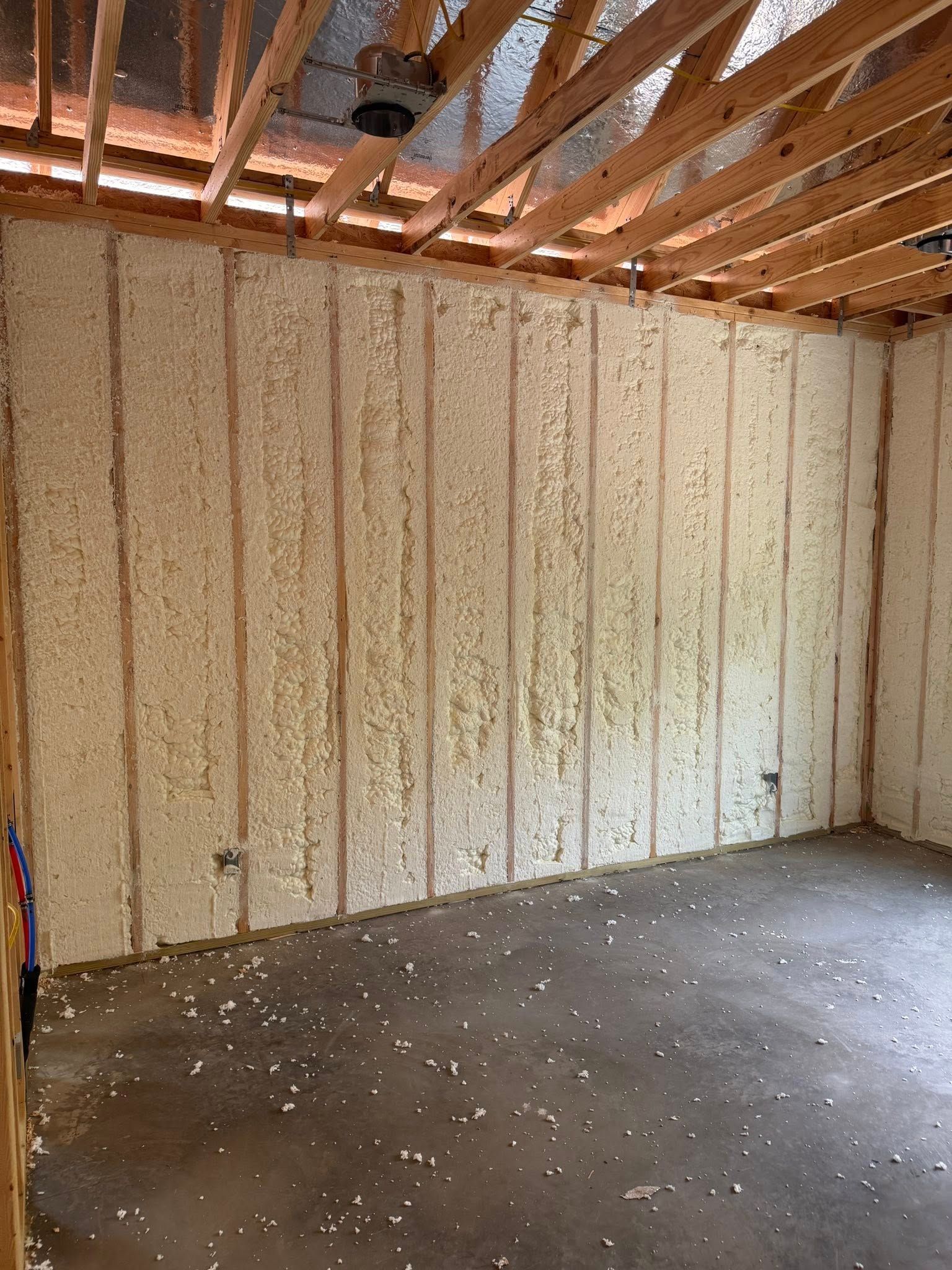 Interior of a room with spray foam insulation on walls, wooden beams, and a concrete floor.