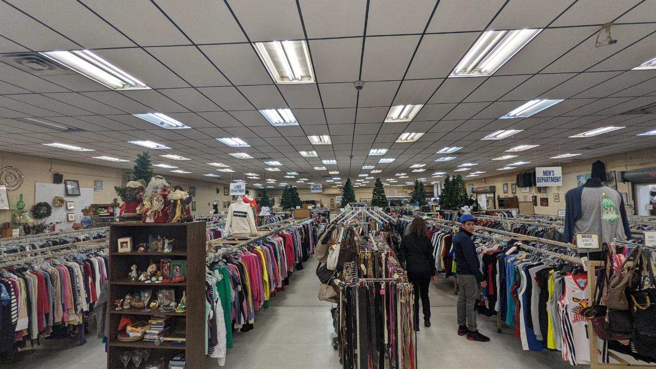 Inside a thrift store; rows of clothing racks, shoppers browsing, overhead fluorescent lighting.